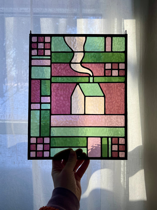 Stained glass panel with geometric textile inspired design with a cozy home depicted in the center, in shades of green and pink, held by a hand in front of softly lit sheer white curtain
