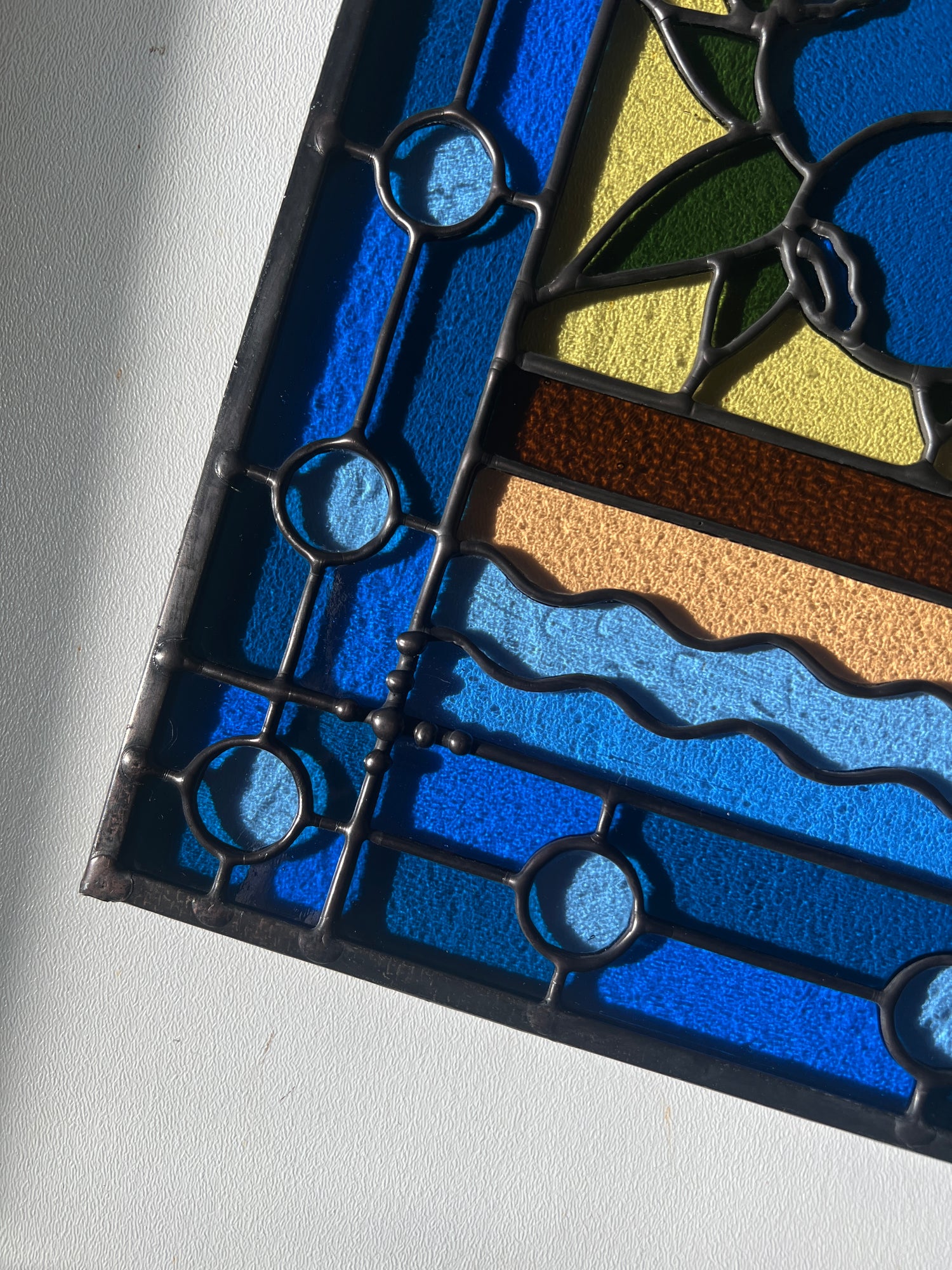 close up of a stained glass panel with a simple compartment style design featuring clouds, sun, water, and a small bunch of blueberries in the middle with a blue border