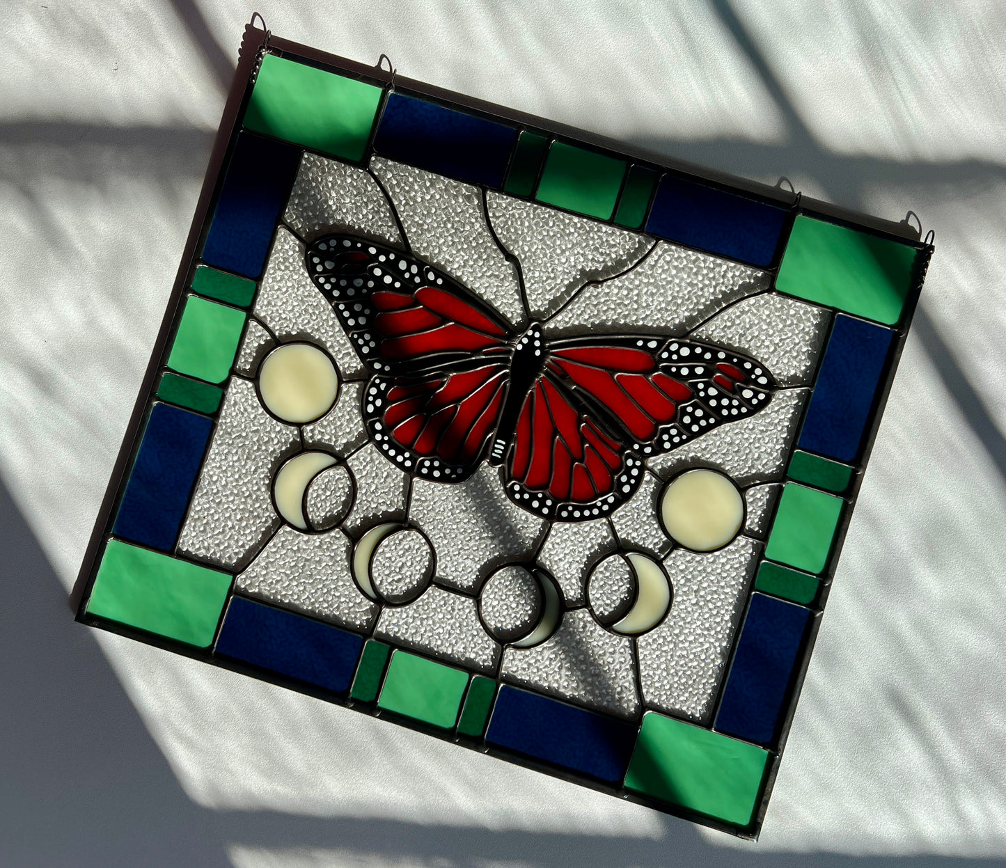 A large stained glass panel with a thick blue and green geometric border and a monarch butterfly and moon phases in the center, with textured clear glass in the background. The piece is laying on a white table with sunlight and shadows falling across it.