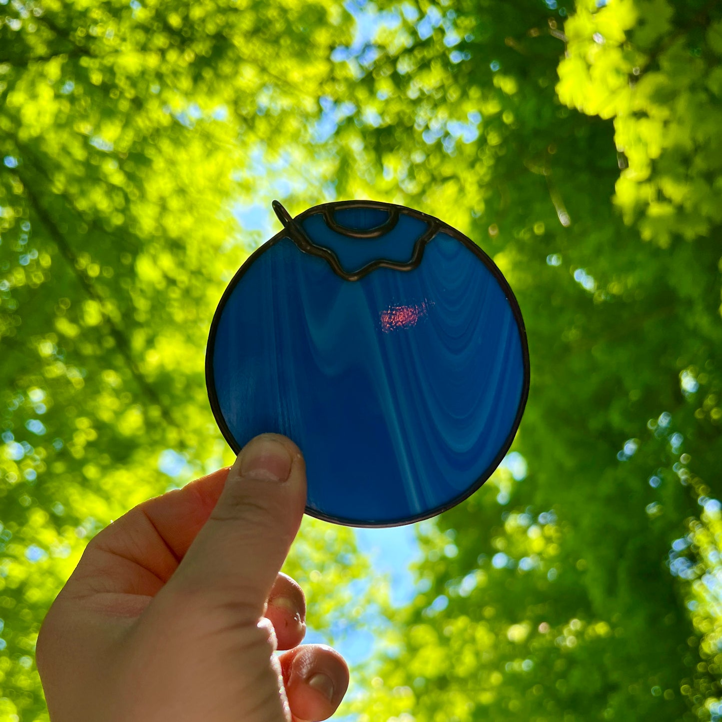 a hand holding a small streaky blue blueberry suncatcher in front of the sun with bright green leaves and trees in the background