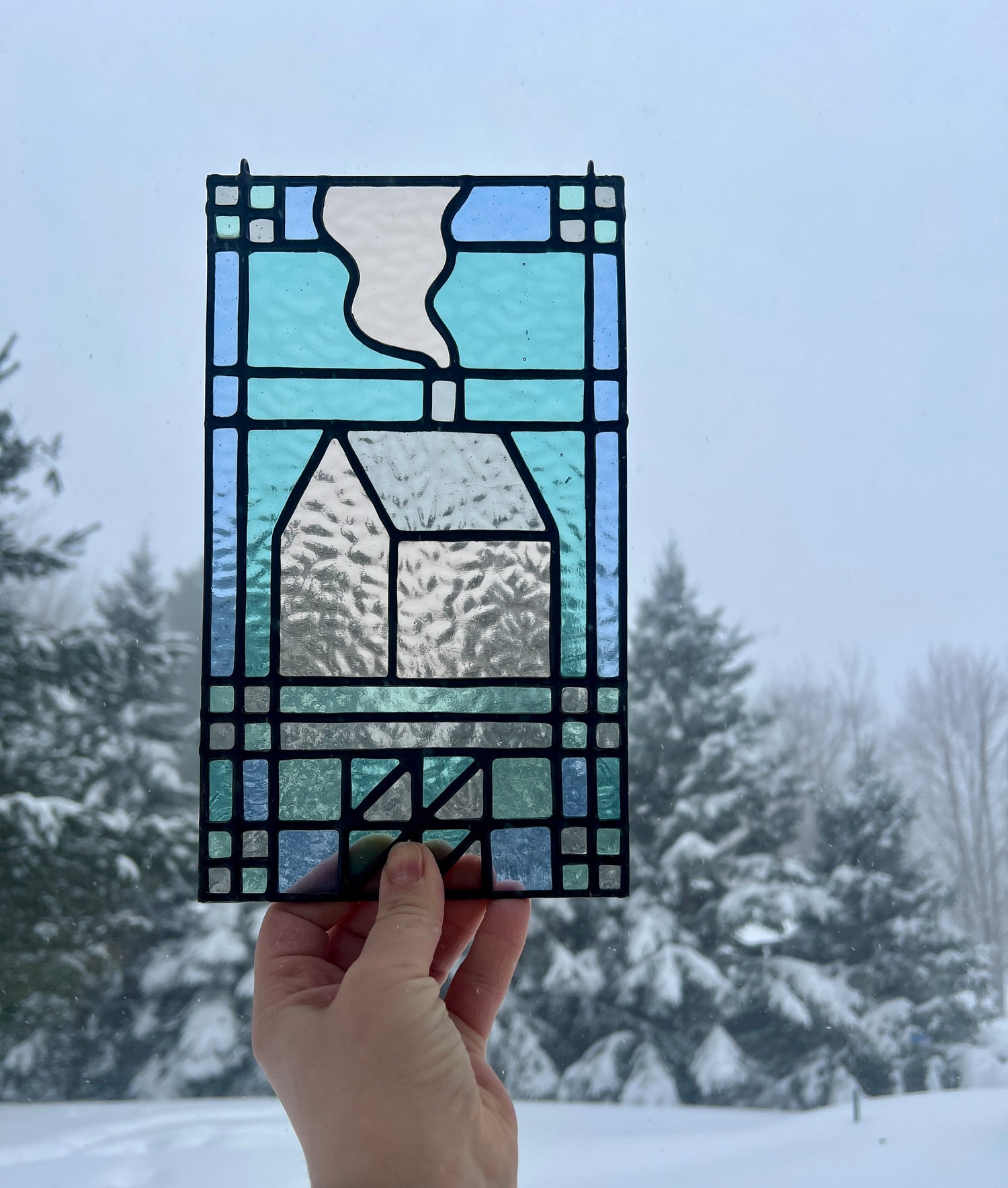 teal and blue stained glass art piece depicting a quilt inspired home, with snow covered pine trees in the background