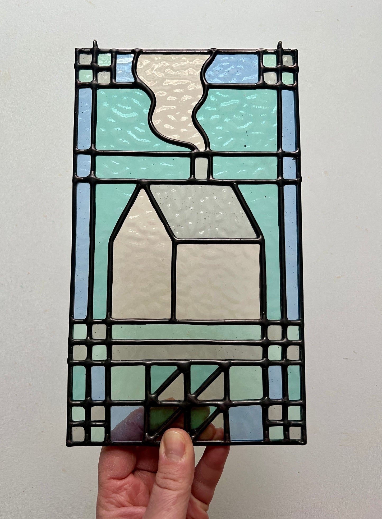 textile inspired cozy home stained glass piece, in pastel shades of blue, teal, and gray, held up by a hand against a white background