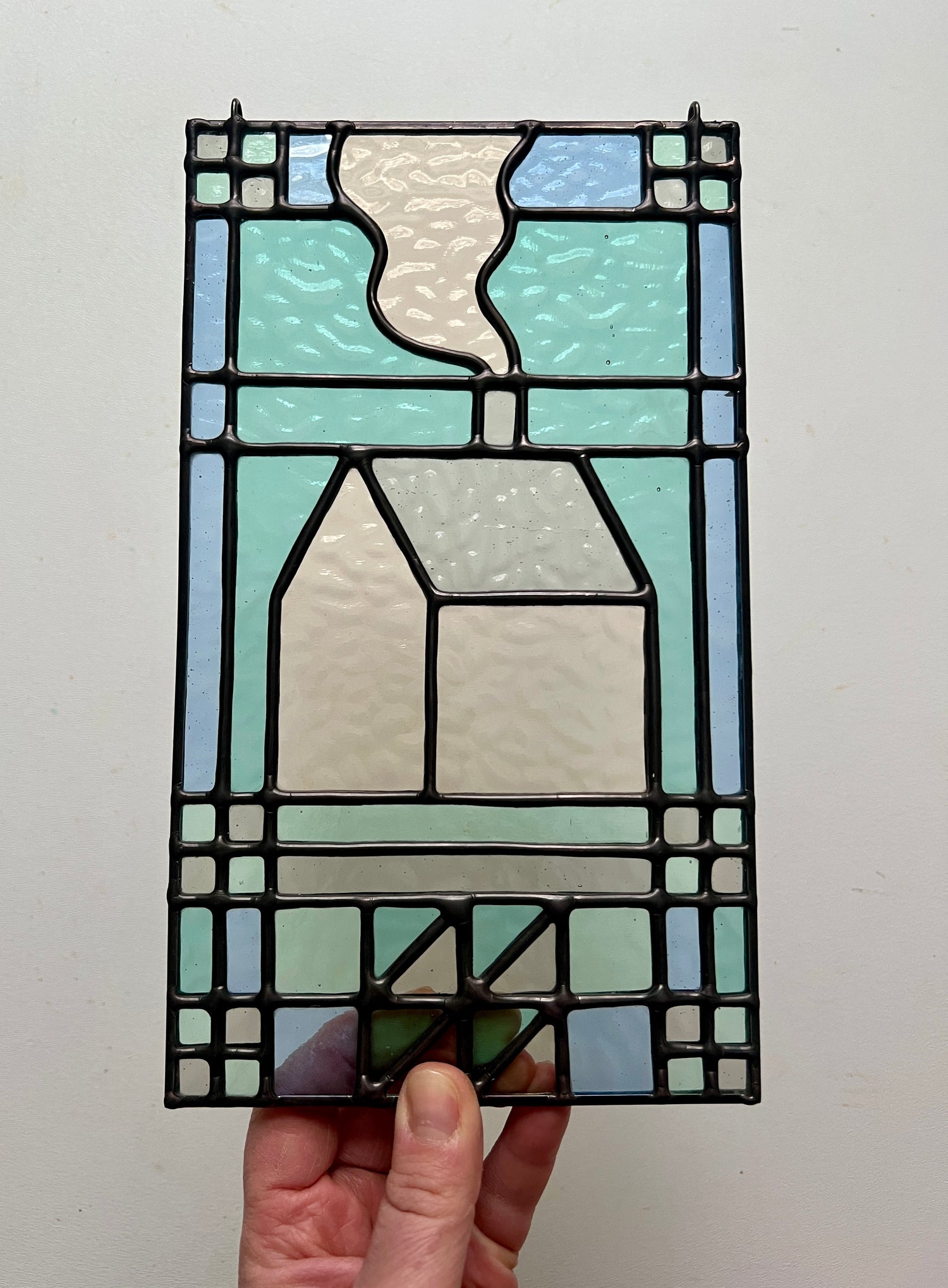textile inspired cozy home stained glass piece, in pastel shades of blue, teal, and gray, held up by a hand against a white background