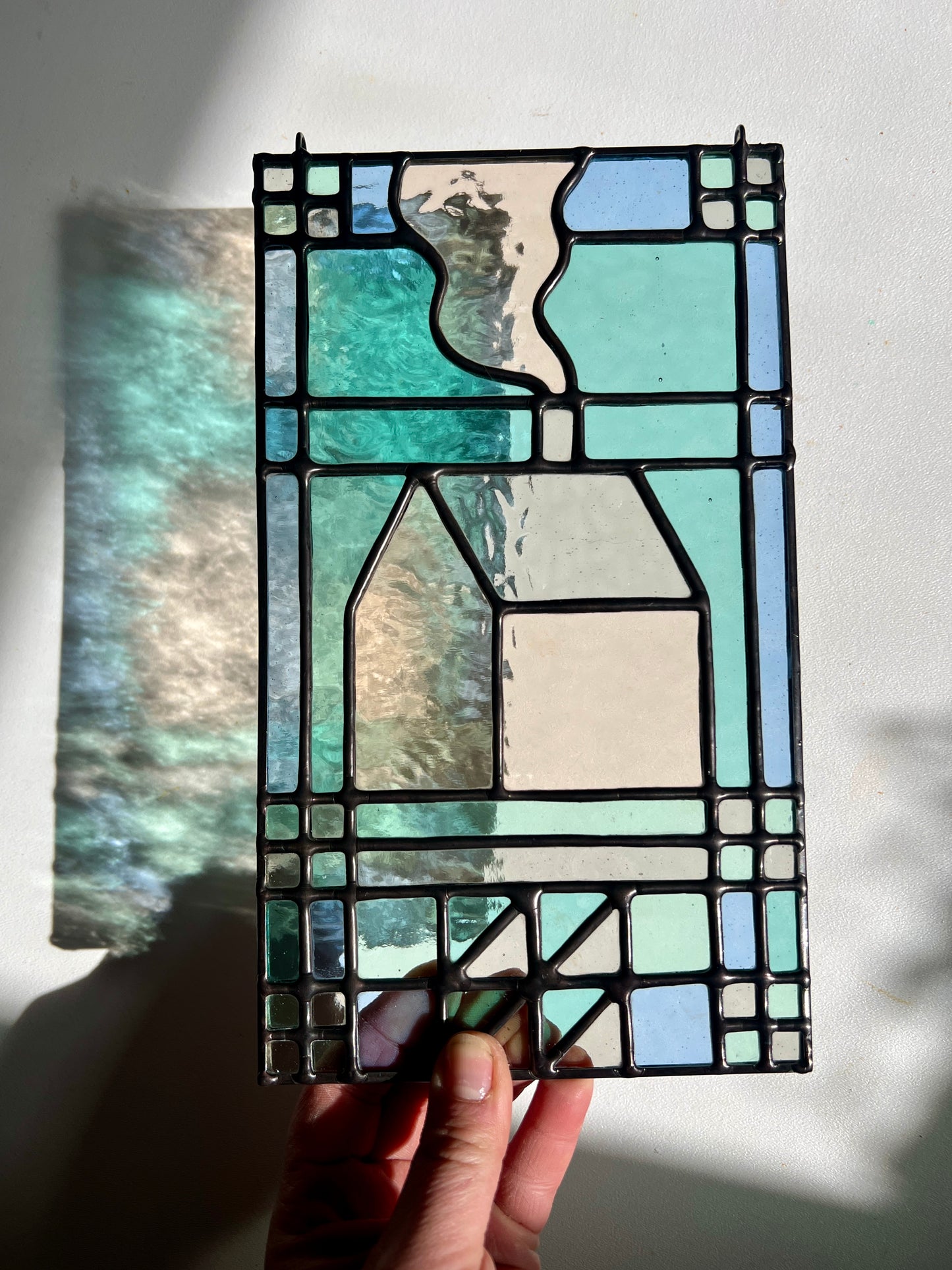 textile inspired cozy home stained glass piece, held by a hand in the sunlight, with soft colorful refractions appearing on the white background
