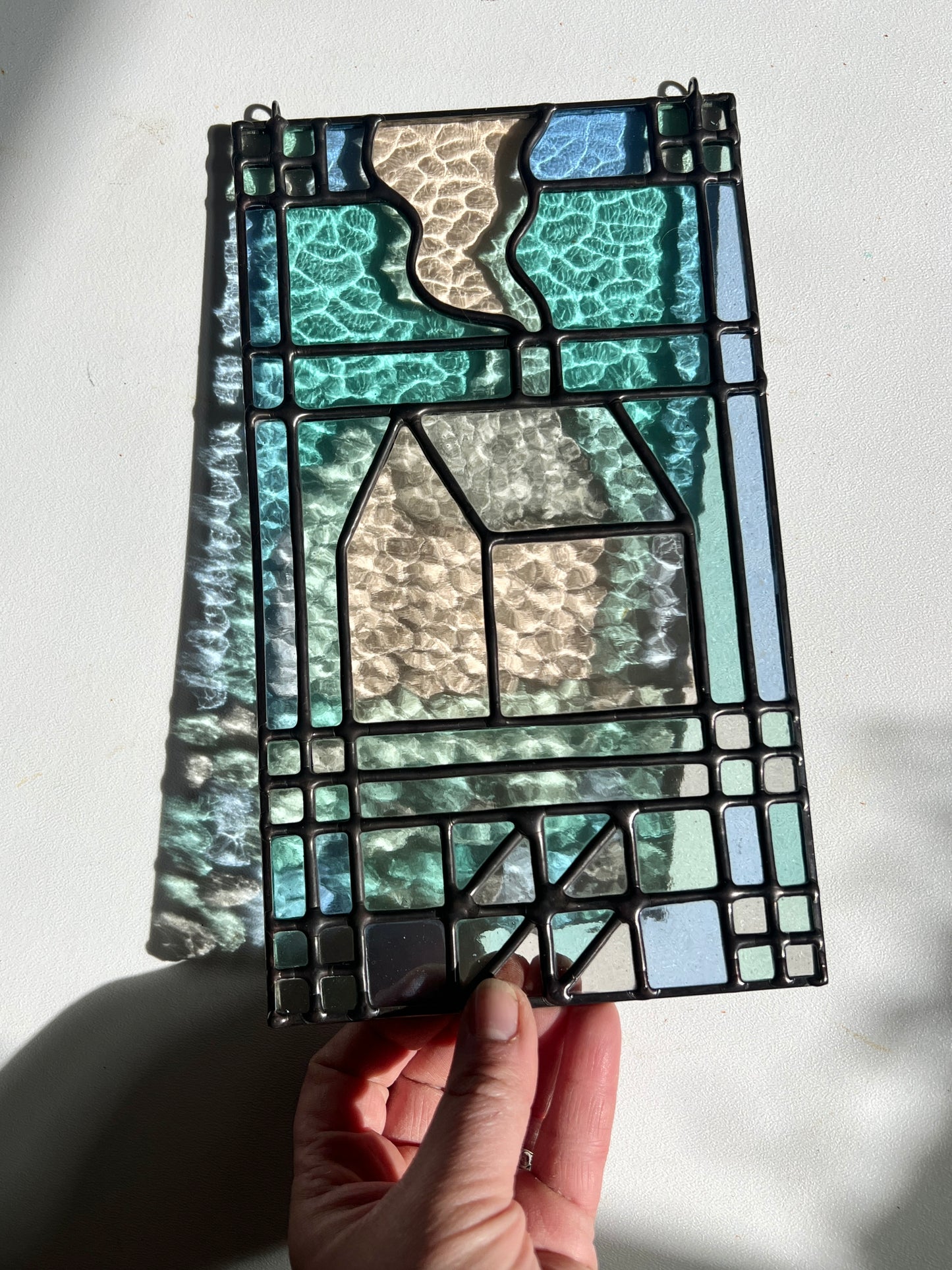 textile inspired cozy home stained glass piece, held by a hand in the sunlight to show the colorful refractions against a white background 